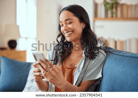 Cheerful latin woman using smartphone during video call while sitting on sofa at home. Smiling hispanic girl chatting on social network on smart phone. Mixed race girl laughing while watching video. Royalty-Free Stock Photo #2357904541
