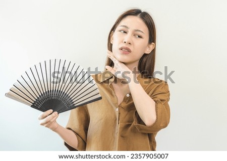 Suffering summer heat stroke, hot weather, tired asian young woman, girl sweaty and thirsty, refreshing with hand in blowing, wave fan to ventilation when temperature high at home, house on background Royalty-Free Stock Photo #2357905007
