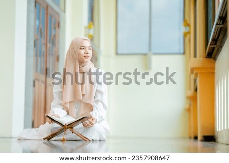 The image of an Asian Muslim woman in the Islamic religion in hijab in cream color. reading the Quran and having a happy Staying in a beautiful mosque, Arabic word translation: The Holy Al Quran.