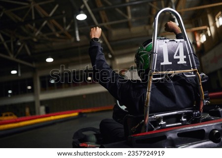 back view of motorsports driver in helmet raising hand and winning race on circuit, go-kart Royalty-Free Stock Photo #2357924919