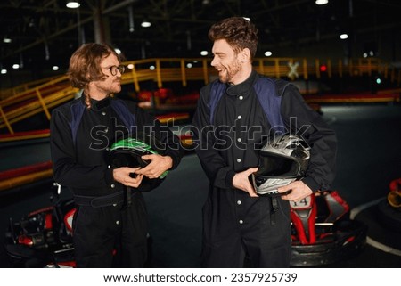 happy two kart racers standing and chatting near racing cars and holding helmets, go-kart drivers Royalty-Free Stock Photo #2357925739