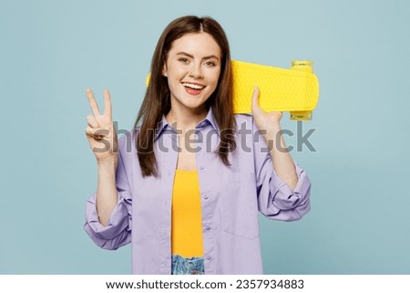 Young happy smiling woman she wears purple shirt yellow t-shirt casual clothes hold skateboard pennyboard show v-sign isolated on plain pastel light blue background studio portrait. Lifestyle concept