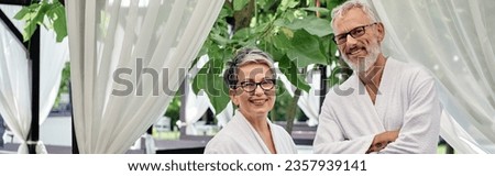 happy middle aged couple in glasses standing in white robes on resort during vacation, banner