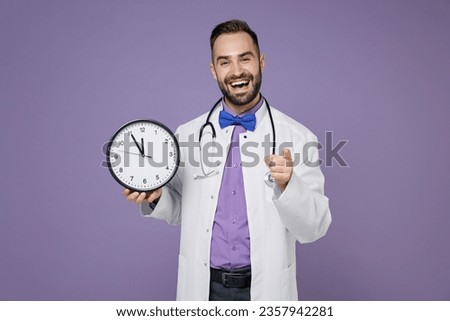 Funny young bearded doctor man in white medical gown stethoscope hold clock pointing index finger on camera isolated on violet background studio portrait. Healthcare personnel health medicine concept
