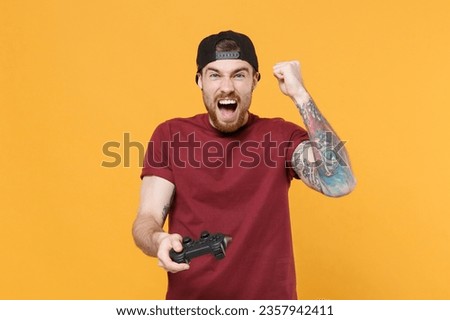 Screaming gambling young tattooed gamer man in casual t-shirt black cap play game with console joystick clenching fist doing winner gesture celebrating isolated on yellow background. Mockup copy space
