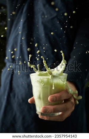 Person holding a glass of matcha green tea which is splashing all around the image frame. Background is dark blue (almost black) apron that male is wearing. Dark vertical exciting image with motion.