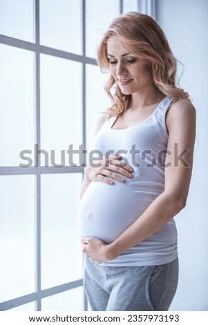 Beautiful pregnant woman is touching her belly and smiling while standing near the window at home
