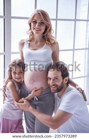 Cute little girl and her handsome dad are hugging her beautiful pregnant mom's tummy, looking at camera and smiling