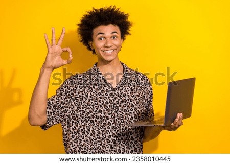 Photo of toothy beaming positive guy with afro hairdo dressed print shirt showing okey holding laptop isolated on yellow color background