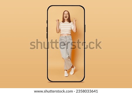 Amazed young woman in casual outfit standing inside huge mobile screen isolated over beige background clenched  fists advertising products winning betting