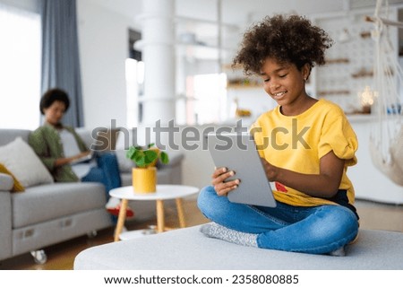 Carefree smiling African American young girl with curly hair using digital tablet sitting with crossed legs at sofa in living room with her mother in background who is using laptop.