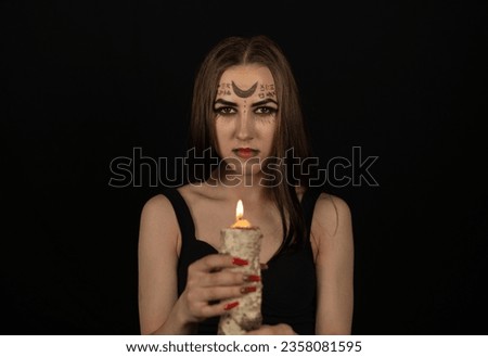 Looking at the camera, a witch with magical symbols on her face holds a burning candle. Halloween. In the dark, a beautiful witch looks askance at the camera. Halloween holiday concept.