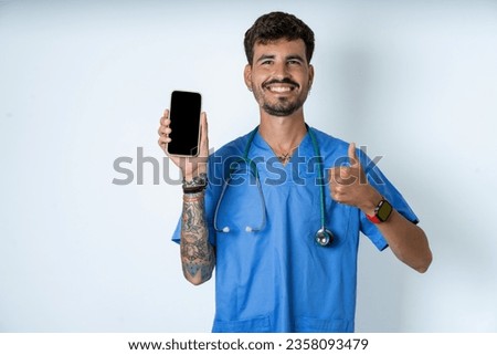 Happy pleased young caucasian doctor man wearing blue medical uniform raises palm and holds cellphone uses high speed internet for text messaging or video calls