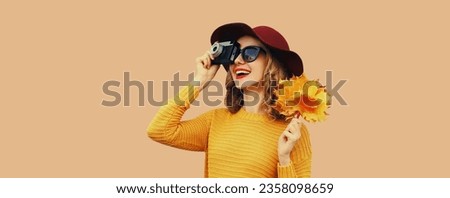 Autumn color style outfit, stylish young woman photographer with film camera holds yellow maple leaves wearing knitted sweater and hat on beige studio background