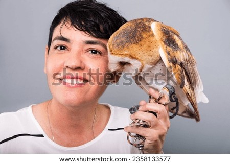 Photo of a woman looking at the camera with her owl next to her face. Wild animal. Studio portrait against gray background.