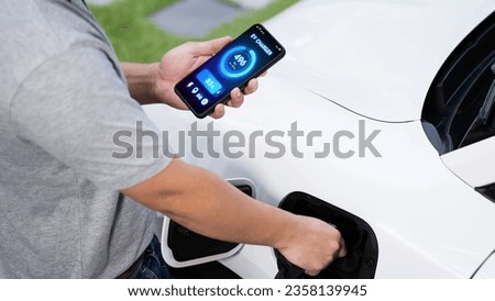 Hand insert EV charger to electric car to recharge battery while check battery status display on smartphone. Future sustainable and clean energy for EV car. Peruse