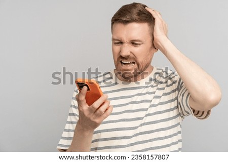 Shocked man clutches head in horror from news from phone, experiencing fail with face distorted grief. Worried male holding smartphone receiving bad tidings, feels like failure, crying from hopeless Royalty-Free Stock Photo #2358157807