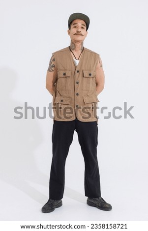 Portrait isolated cutout studio full body shot Asian vintage classy mustache neck arms tattoos male fashion model in casual fashionable safari vest and cap standing look at camera on white background.