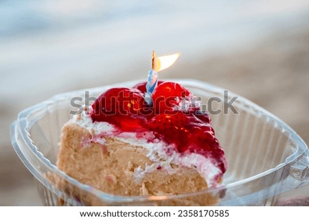 Bright close-up picture of slice of strawberry cheescake with blue birthday candle inside plastic container on the beach