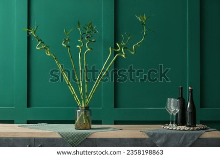 Vase with bamboo stems, bottles of wine and glasses on table near color wall
