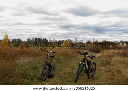 Two bicycles standing in autumn field, meadow with brown grass. Fall country landscape with brown and orange forest trees, dried grass, sky with gray clouds. Travel cycling break, destination point