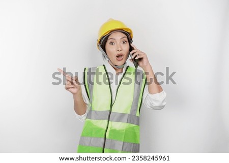 A smiling Asian woman labor wearing safety helmet and vest, pointing to copy space beside her while having a phone call, isolated by white background. Labor's day concept.