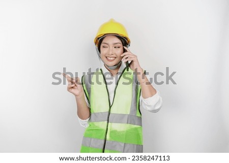 A smiling Asian woman labor wearing safety helmet and vest, having a phone call and pointing to copy space beside her, isolated by white background. Labor's day concept.