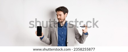 Online shopping. Smiling business man in suit showing plastic credit card with empty smartphone screen, standing against white background.