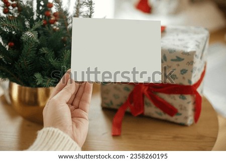 Christmas card mock up. Hand holding empty greeting card on stylish wrapped christmas gift and fir branches with festive christmas lights on wooden table. Space for text. Season greetings postcard