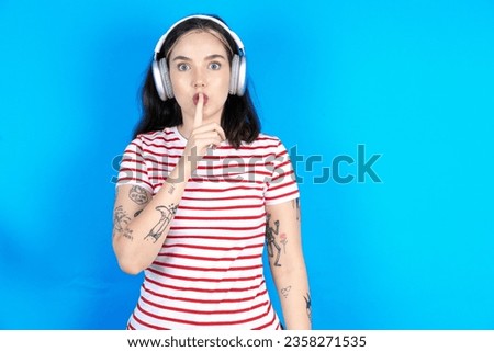 Young caucasian woman wearing striped t-shirt making hush gesture with finger on her lips wearing  wireless headphones. Be quiet.