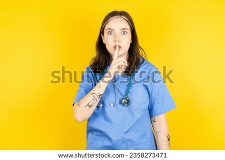 Surprised Young caucasian doctor woman wearing blue medical uniform makes silence gesture, keeps finger over lips and looks mysterious at camera