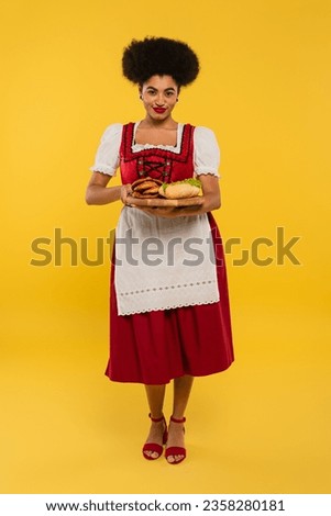 pleased african american bavarian waitress serving pretzels and hot dog on wooden tray on yellow