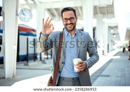 Man with drinking a cup of coffee doing ok sign with fingers, excellent symbol. Young smiling bearded drinking hold paper cup of coffee show thumb up gesture on train station. 