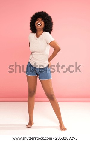 Optimistic african american woman having fun , smiling, laughing happily. Girl with afro hairstyle wearing casual tshirt and short jeans, posing on pink pastel background. 

