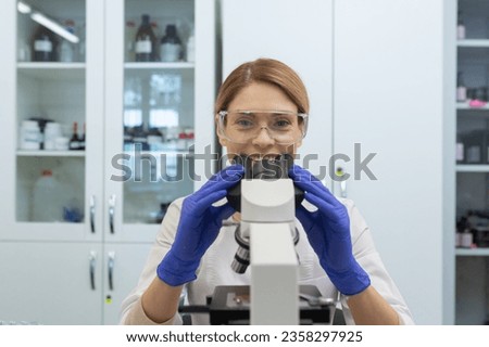 Close-up photo of a young woman working in a laboratory under a microscope, sitting at a table in a uniform and glasses and smiling at the camera.