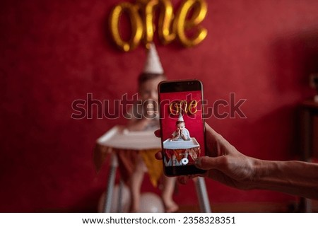 Mothers hand takes pictures of son on a smartphone. A little boy of one year in festive hat holding birthday cake with a candle in hands. On red plain background gold foil balloons One. Social Media