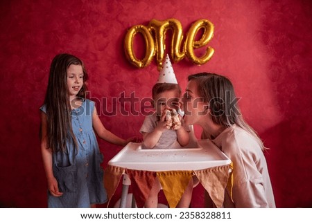 Older sister and mother help little boy blow out candle for his first birthday. Children with young woman have fun on red background with foil balloons. Home party with meringue cake. Single parent