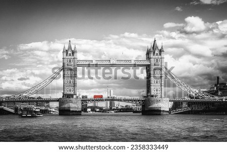 An iconic red London bus highlighted in a black and white image as it passes over Tower Bridge on the river Thames in London, England