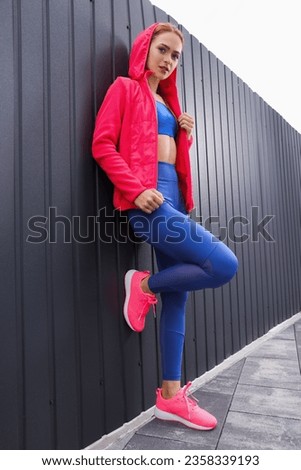 Beautiful woman in gym clothes posing near dark grey fence on street, low angle view