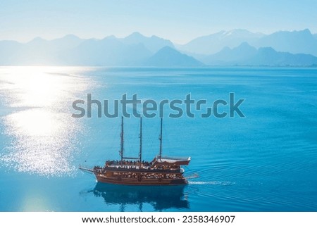 Historic wooden ship with sails floats on the water of the sea surface, a bay surrounded by mountain peaks with a bright sun, aerial view