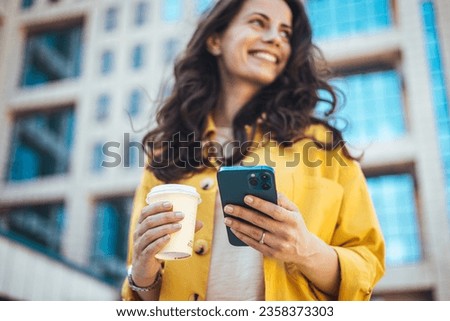 Woman using smartphone and drinking coffee at the city. Closeup shot of a businesswoman using a cellphone in the city. Young smiling woman holding a smartphone and a cup of coffee