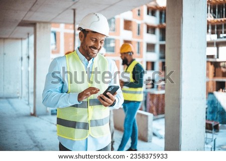 Shot of a young businessman using his smartphone while on a construction site. Portrait of young engineer in vest with white helmet standing on construction site, smiling and holding smartphone 