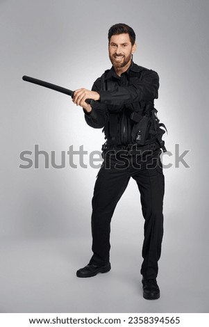 Aggressive dark haired police officer brandishing with police baton in studio. Front view of angry, bearded man in police uniform beating, attacking camera, on gray background. Concept of emotions.