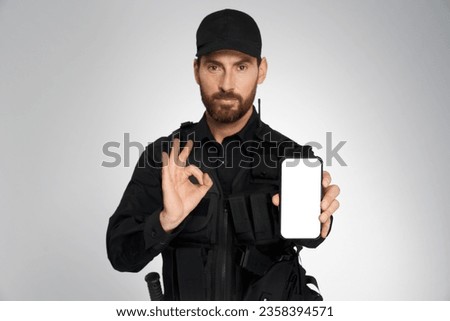 Serious male police officer showing empty mobile phone's screen, with copy place. Portrait of bearded policeman gesturing ok sign, looking camera, isolated on gray. Concept of safety, phone call. Royalty-Free Stock Photo #2358394571