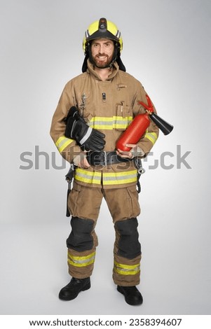Young firefighter in helmet holding fire extinguisher in studio. Front view of bearded man wearing fireman uniform, smiling, isolated on white background. Concept of safety, work, rescue.