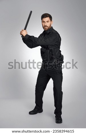 Focused caucasian police officer brandishing with police baton. Front view of dark-haired guy in police uniform beating, attacking camera, on gray background. Concept of police work, equipment.