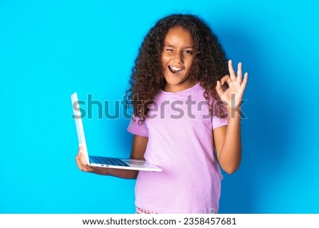 Attractive cheerful skilled Beautiful kid girl standing over blue background using laptop showing ok-sign winkin