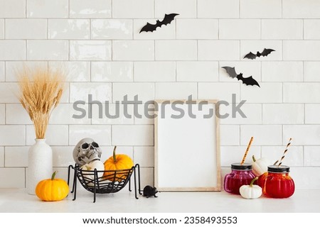 Halloween holiday concept. Empty picture frame mockup, vase of dried flowers, drinks, pumpkins, skull on white brick wall background.