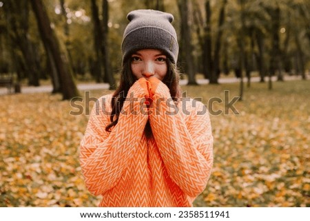 Caucasian woman walking outdoor, portrait of young european lady in warm sunny autumn park season, fall, yellow orange red leaves, feeling cold, rubbing hands blowing on palms
 Royalty-Free Stock Photo #2358511941
