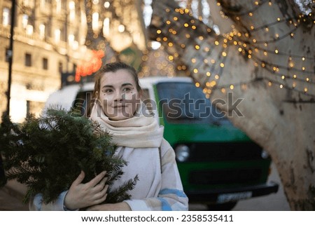 Christmas photo. Young 30-year-old beautiful brunette childfreewoman at Christmas market holds fir branch in her hand. New Year gift shopping concept. Retro car on background. Bokeh lights. Garland